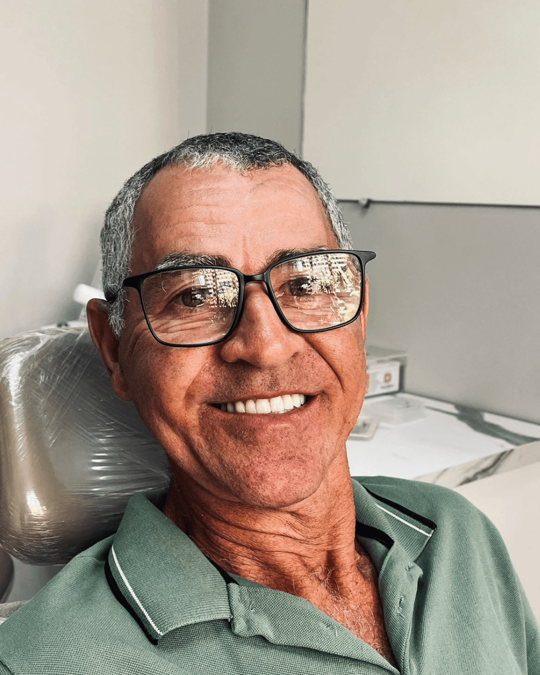 Paciente homem de óculos e camisa verde sorri na cadeira clínica após reabilitação com implante dentário e prótese protocolo em Franca SP.