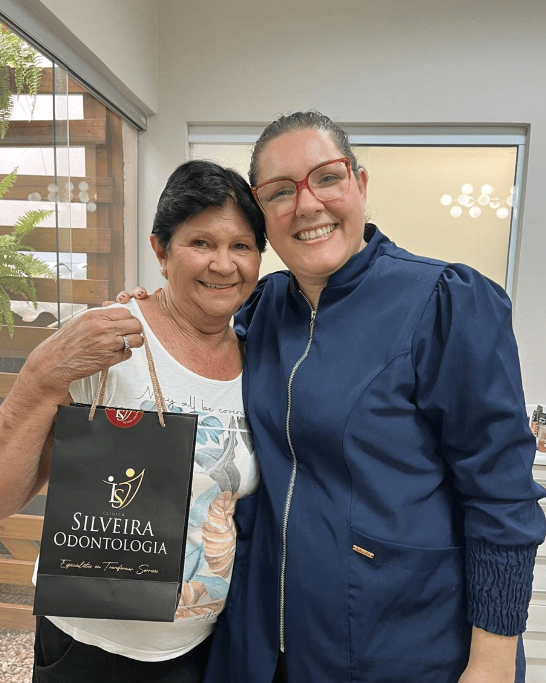 Dentista de jaleco azul abraça paciente que segura sacola da Silveira Odontologia após finalizar tratamento de implantes em Franca SP.