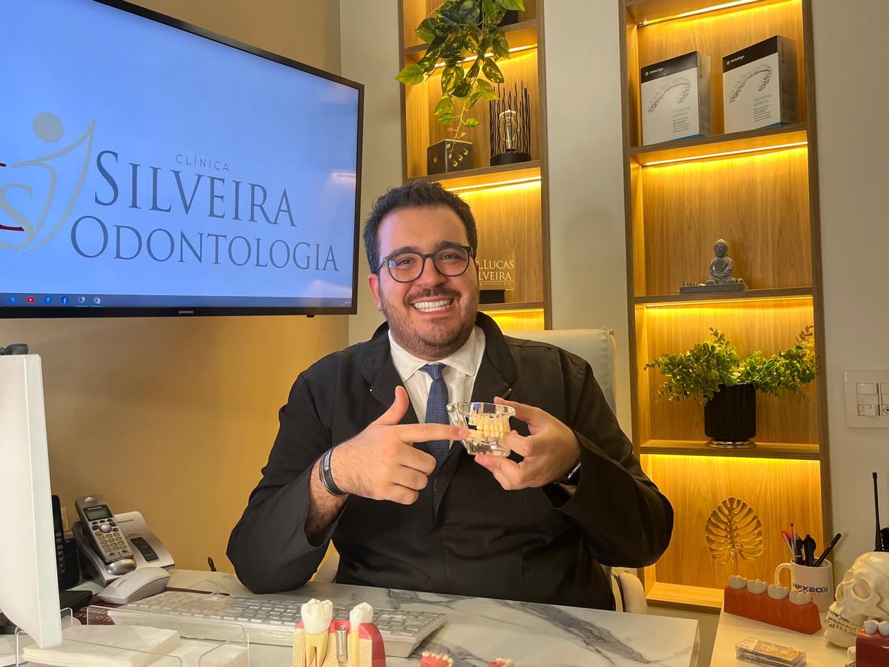 Dentista sorrindo aponta para modelo anatômico de arcada dentária na Silveira Odontologia, Clínica Odontológica em Franca, explicando implantes dentários em Franca SP. Especialistas em Cirurgia Odontológica Bucomaxilofacial em Franca sp